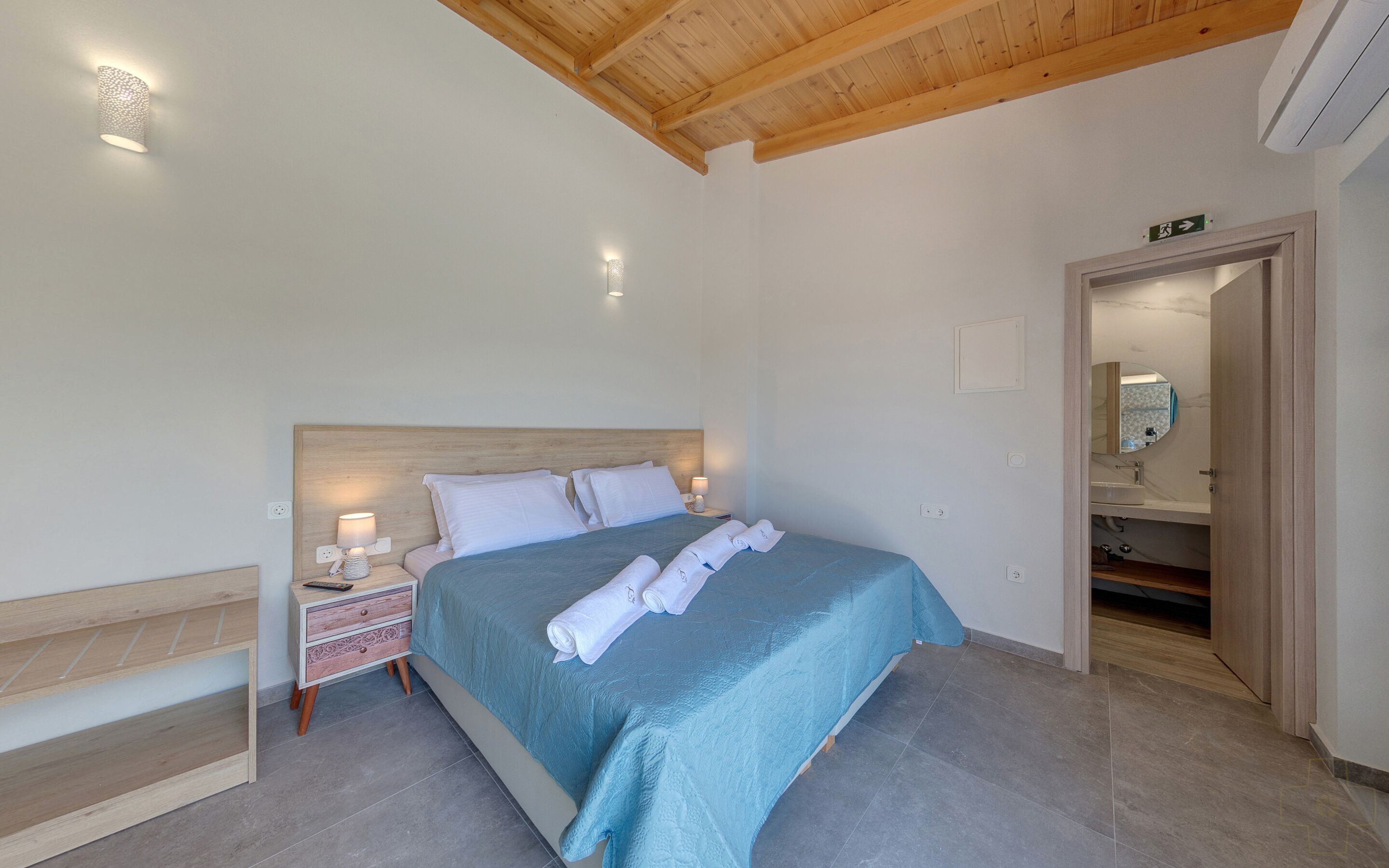 Un dormitorio con cama, mesitas de noche, cabecera de madera y techo con vigas de madera, además de una puerta abierta que da a un baño.