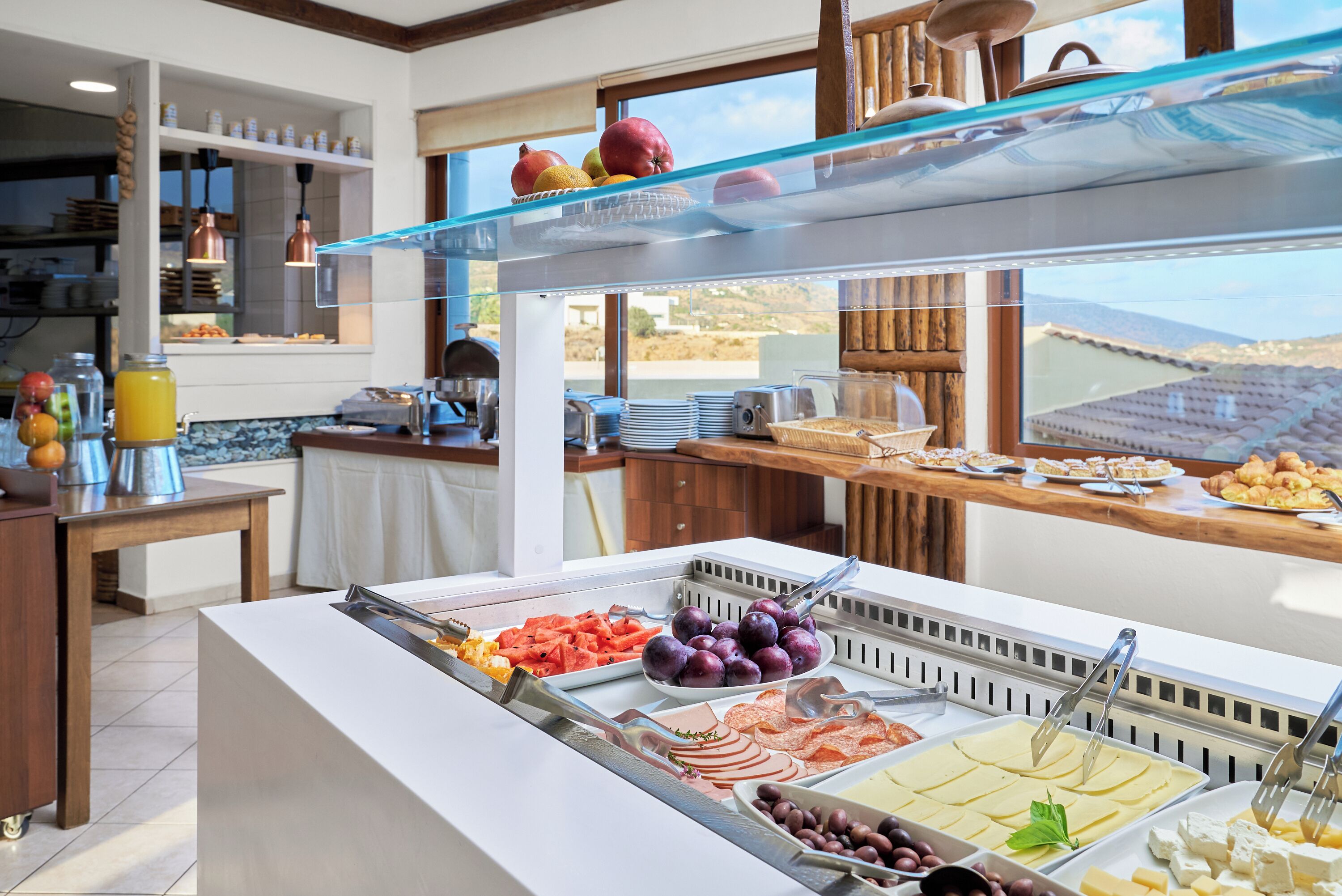 A buffet with various food items, including fruits, cheeses, and meats, in a well-lit room with a view of the outdoors.