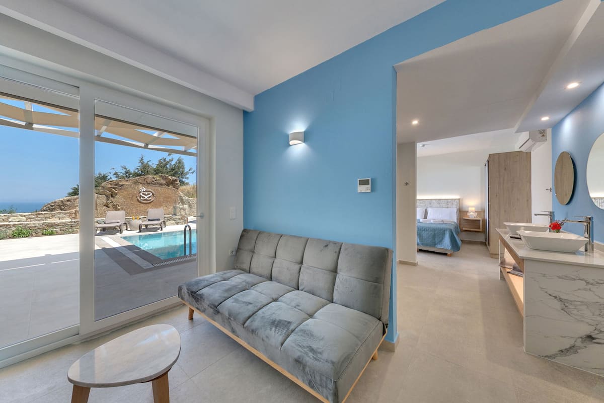 Une chambre à coucher moderne, dotée d’un grand lit, d’une salle de bain avec un plan de toilette en marbre et offrant une vue sur la mer grâce à une porte vitrée.