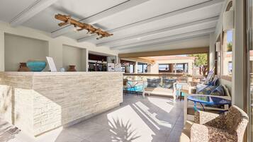 A sunlit outdoor seating area with a stone reception desk, cushioned seating, and a pool view.