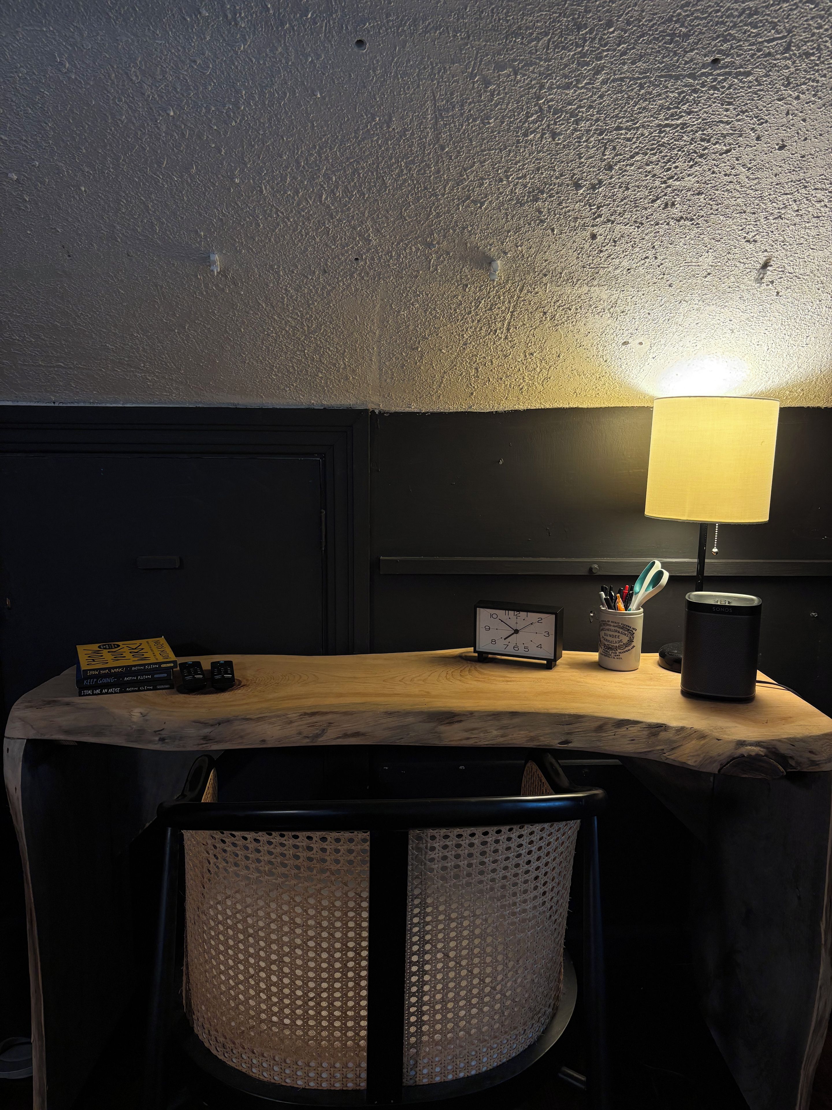 A small, dark room with a wooden table, a lamp, a clock, and a speaker.