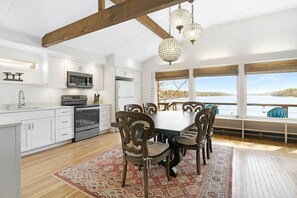 A modern kitchen with white cabinets, a stainless steel oven, and a microwave. There is a dining table with chairs and a view of a lake through large windows.