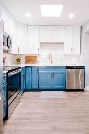 A modern kitchen with white upper cabinets, blue lower cabinets, stainless steel appliances, and a light wood floor.