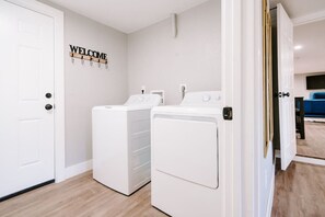 A laundry room with two white washing machines, a 'WELCOME' sign on the wall, and a blue couch in the background.