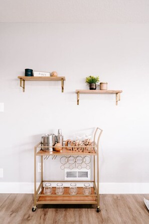 Ein minimalistischer Raum mit zwei Wandregalen aus Holz, auf denen dekorative Gegenstände und je eine kleine Pflanze stehen. Darunter ein Metallwagen mit Glasbehältern und einer Metallkanne auf Holzdielen.