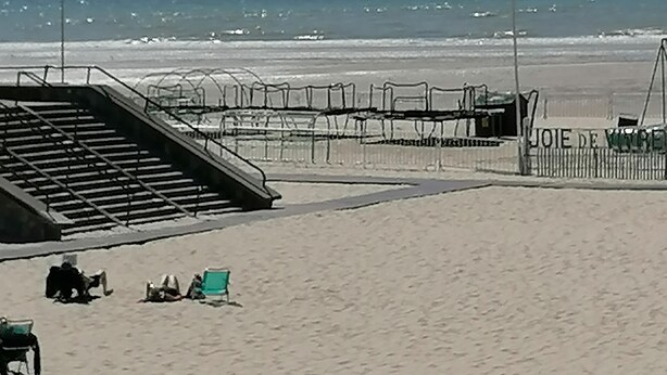 Una playa con una escalera que baja hacia la arena, una cerca con un cartel y algunas sillas.