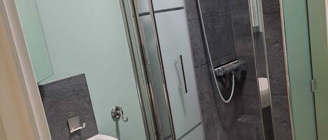 Un baño moderno con ducha de vidrio, lavamanos blanco y paredes de azulejos oscuros.
