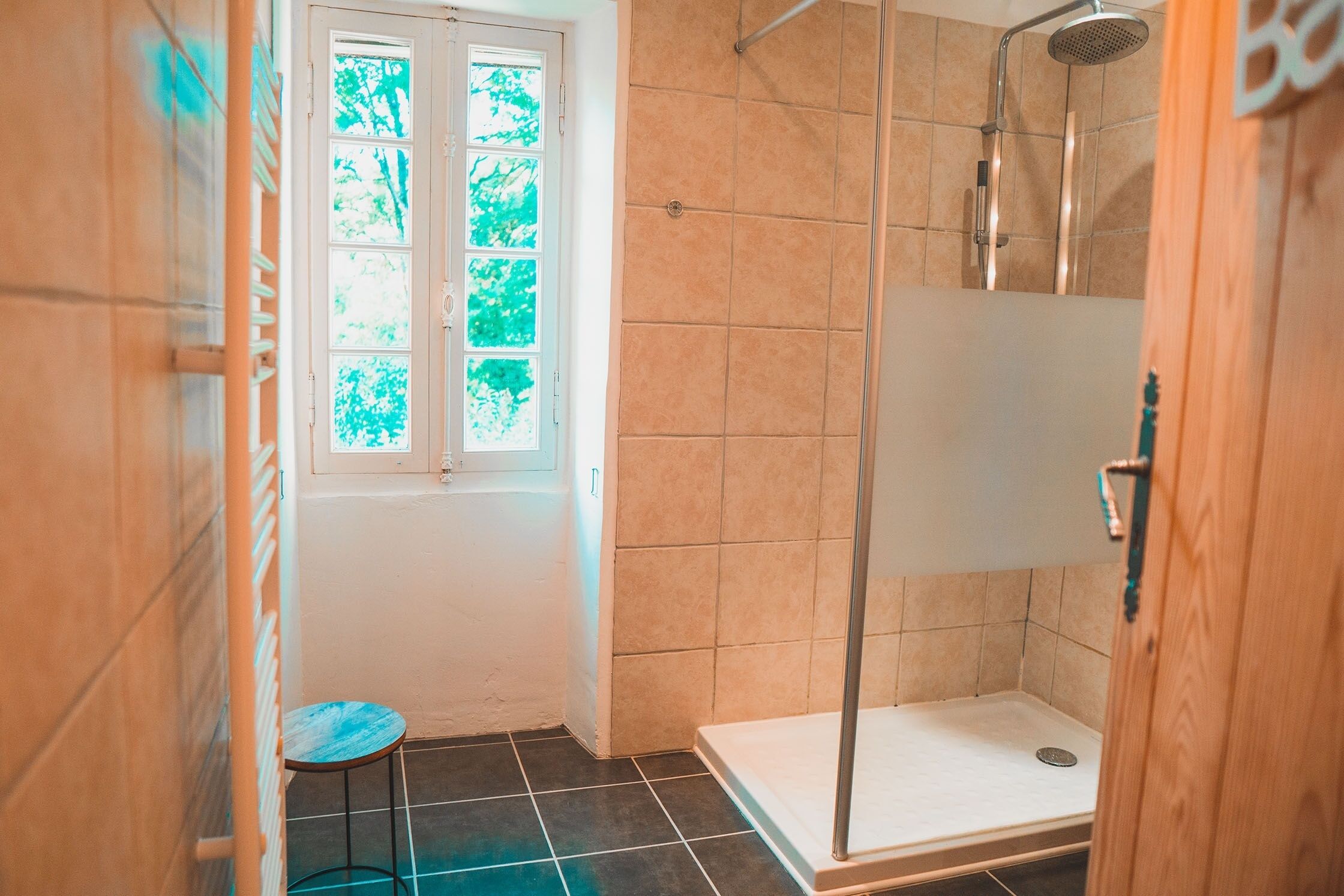 A shower with a glass door, a small stool, and a window with a view of trees.