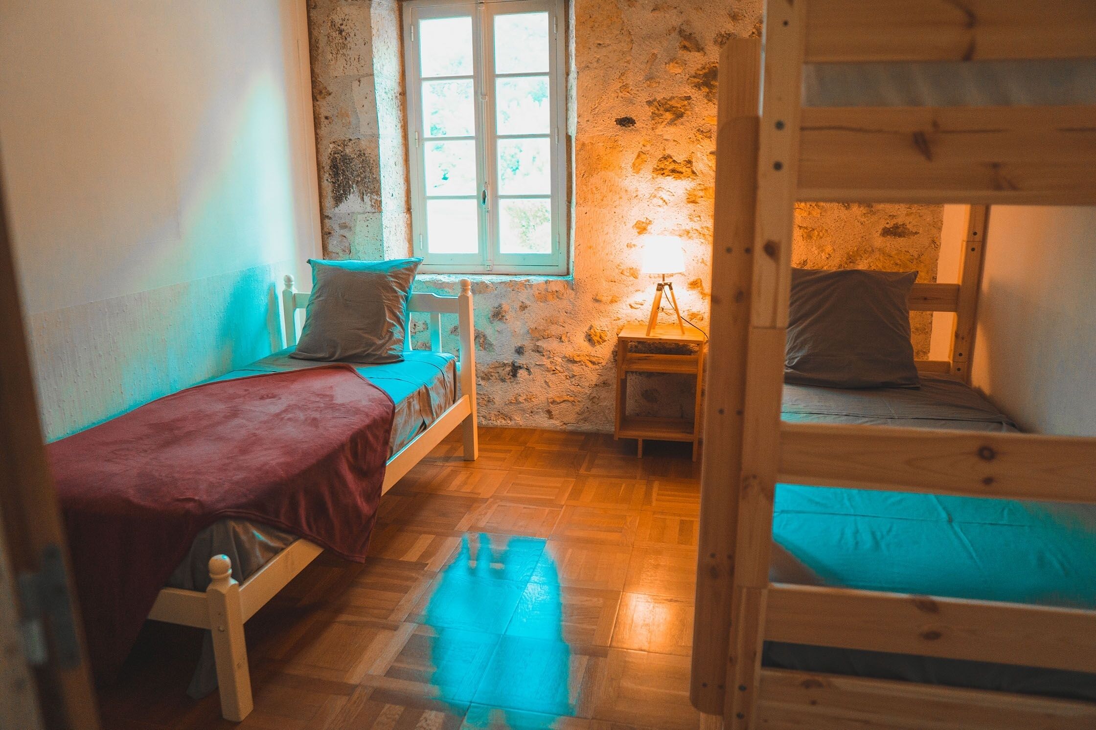 A dormitory room with bunk beds, a single bed, a window, and a bedside lamp.