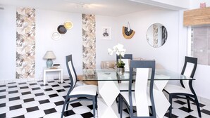 En matplats med ett glasbord, svarta och vita stolar, ett litet sidobord med en lampa och en vägg dekorerad med prydnadstallrikar och en spegel.
