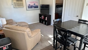 A living room with a beige sofa, a TV on a stand, a dining table with chairs, a bookshelf, and a door leading to another room.