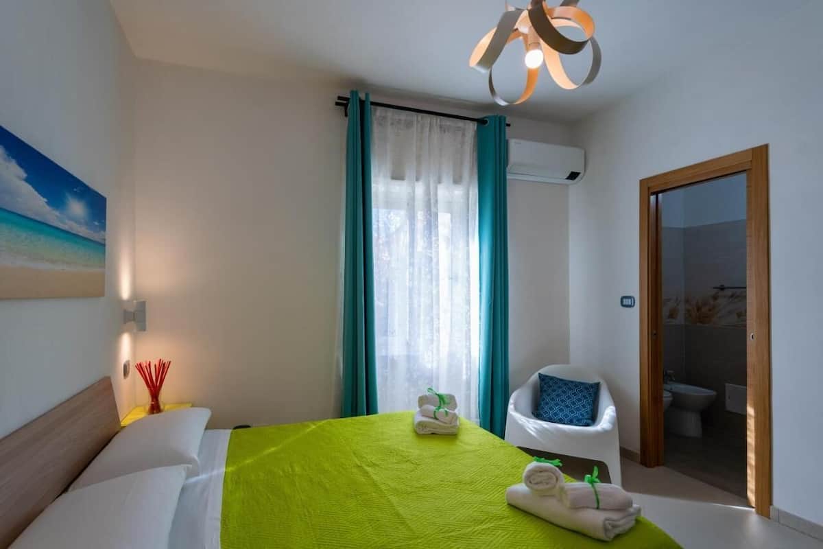 A hotel room with a bed, a chair, a bathroom visible through an open door, and a painting on the wall.