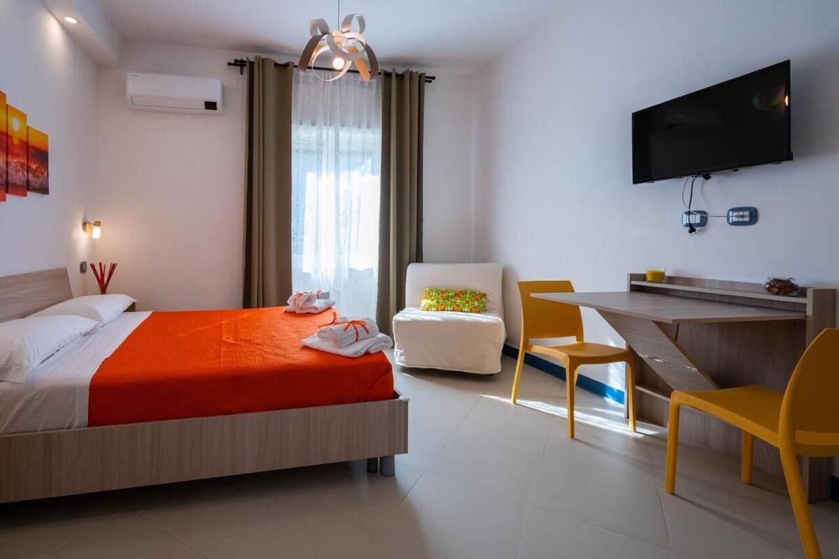 A hotel room with a bed, a desk, two chairs, a TV, and an air conditioning unit.