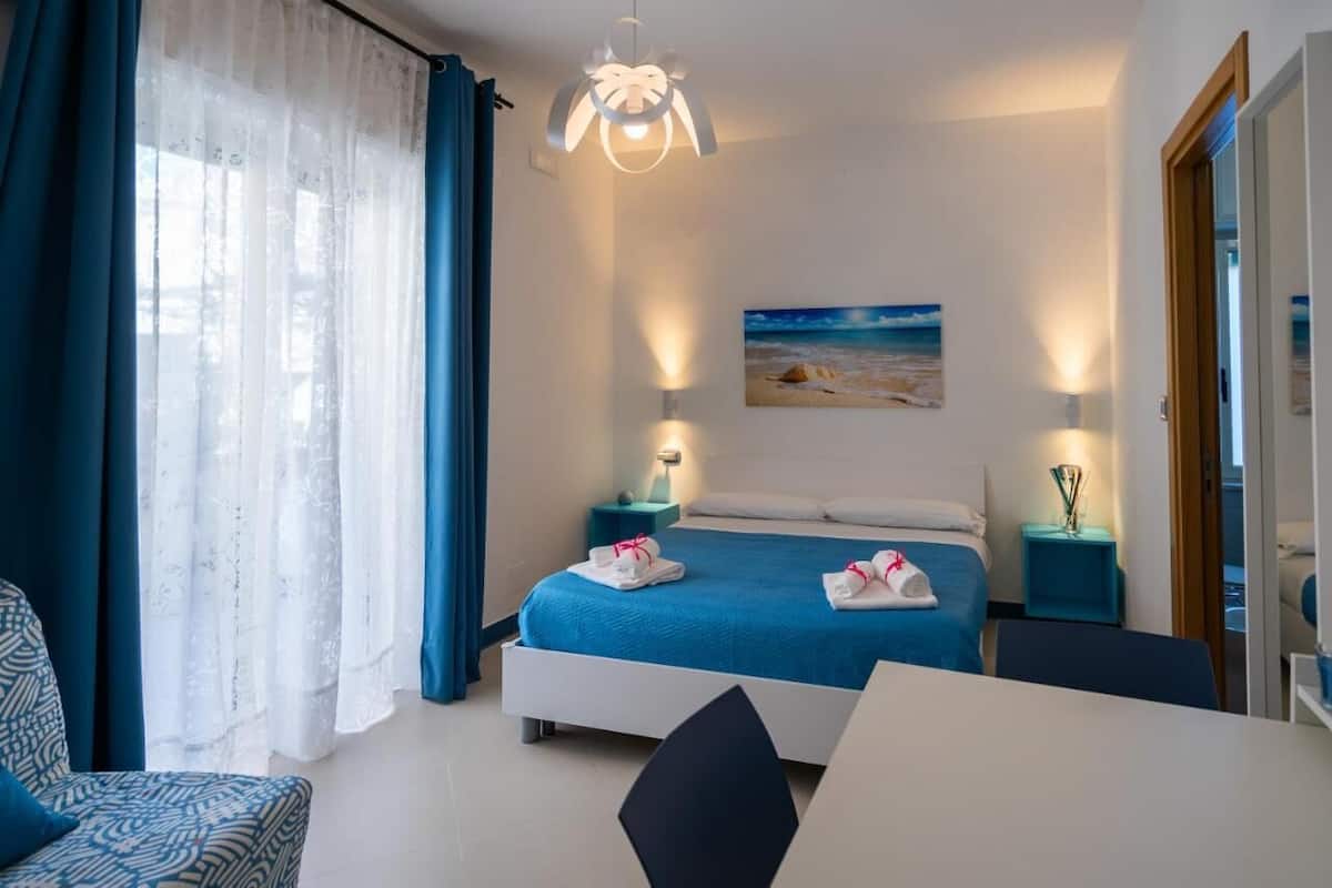 A hotel room with a bed, two bedside tables with lamps, a painting on the wall, and a view of the beach.
