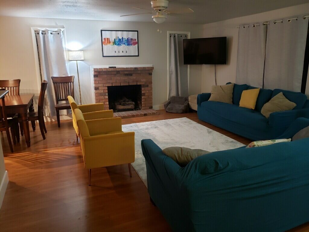 Een woonkamer met een open haard, een blauwe bank, gele fauteuils en een aan de muur bevestigde televisie.