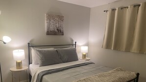 Ein ordentlich bezogenes Bett mit Kopfteil, zwei Nachttischlampen, einem Wandbild und einem Fenster mit Vorhängen.