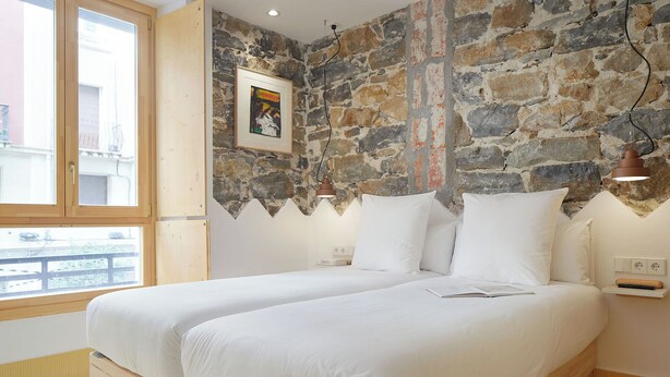 Une chambre avec un mur en pierre, deux lits, une fenêtre et une lampe.