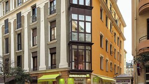Un immeuble d’angle à plusieurs étages, une devanture de magasin avec un auvent vert et un trottoir pour les piétons.