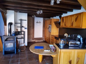 A rustic kitchen with a wood stove, wooden cabinets, a round table, and a hanging light fixture. - Le Dorli Schopf Gîte (Dorlisheim)