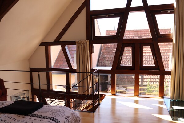 A room with a sloped ceiling, wooden beams, a bed with patterned bedding, a window with curtains, and a view of buildings outside.