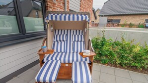 A sun lounger with striped cushions and wooden legs, positioned on a tiled patio with a brick wall and a window in the background.
