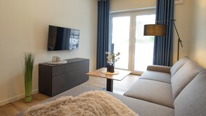 A modern living room with a sofa, a coffee table, a TV mounted on the wall, and a large window with curtains.