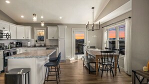 A modern kitchen with white cabinets, a large island, and a dining area with a view of the sunset.