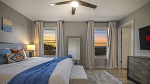 A bedroom with a bed, bedside table, lamp, and a view of the sunset through large windows.