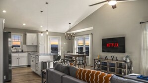 A modern kitchen with white cabinets, a dining area with a table and chairs, a gray sofa with a patterned throw, and a TV mounted on the wall displaying "NETFLIX".