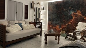 Ein modernes Wohnzimmer mit einer Couch, einem Sessel und einem Couchtisch. An der Wand befindet sich ein großes Wandbild, Regale mit Dekorationselementen und ein Fenster mit Fensterläden.