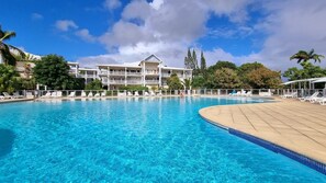 Une piscine d’un bleu éclatant, bordée de chaises longues blanches, un immeuble à plusieurs étages doté de balcons, et une végétation luxuriante en arrière-plan.