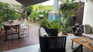 Eine Terrasse mit Holztisch, Stühlen, einer Couch und einem Couchtisch. Übertöpfe mit Pflanzen und ein dekoratives Steinobjekt sind ebenfalls vorhanden.