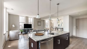 Una cocina moderna con una isla central, electrodomésticos de acero inoxidable, iluminación colgante y vistas al salón.