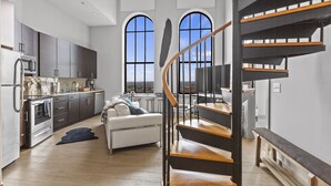 A modern kitchen with dark cabinets, a white countertop, and a built-in oven. There is a white sofa with a blue throw blanket in the living area. A wooden staircase with a black railing leads to an upper level. - Distinguished Apartment Rooftop Deck (Baltimore)