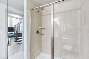 A modern bathroom with a glass shower enclosure, a white tiled floor, and a visible staircase leading to another area. - Distinguished Apartment Rooftop Deck (Baltimore)