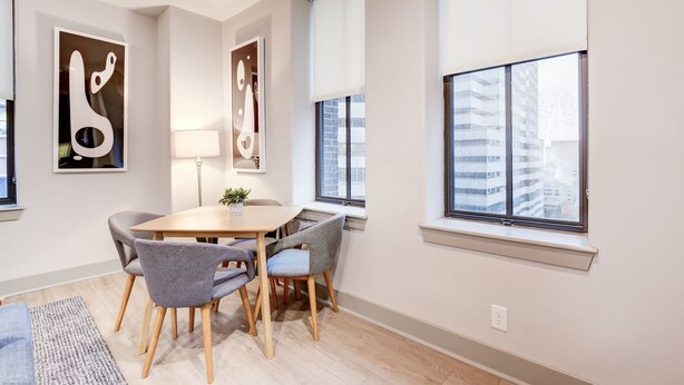 A modern dining area with a round table, four grey chairs, and a small plant. There are two large windows with blinds, and a wall-mounted lamp. - Distinguished Apartment Rooftop Deck (Baltimore)
