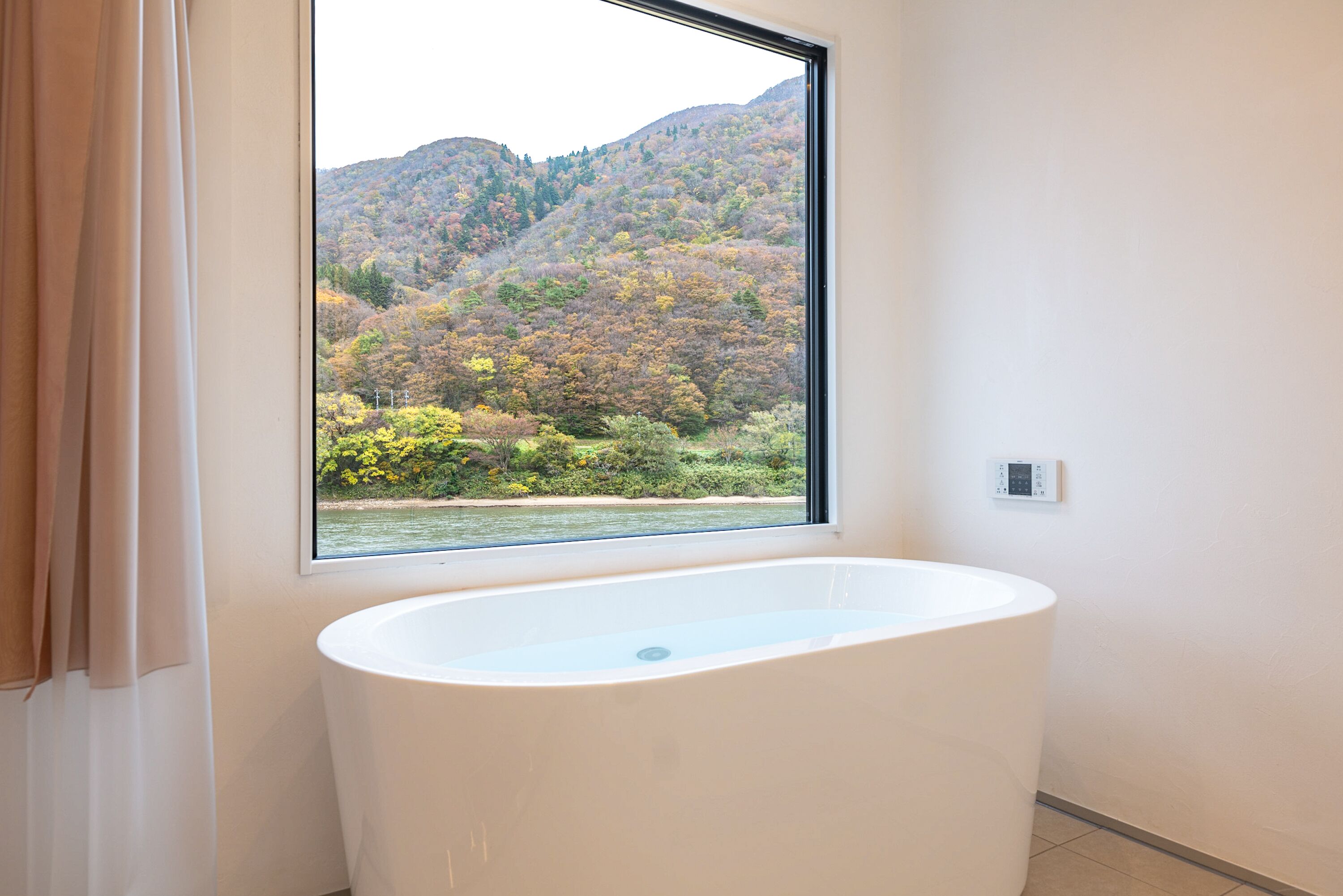 A bathroom with a bathtub, a window with a view of a forested hillside, and a thermostat on the wall.