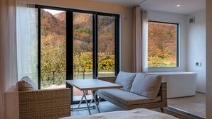 A bedroom with a bed, a small table, a sofa, and large windows offering a view of autumn foliage. - R MOGAMI (Tozawa)