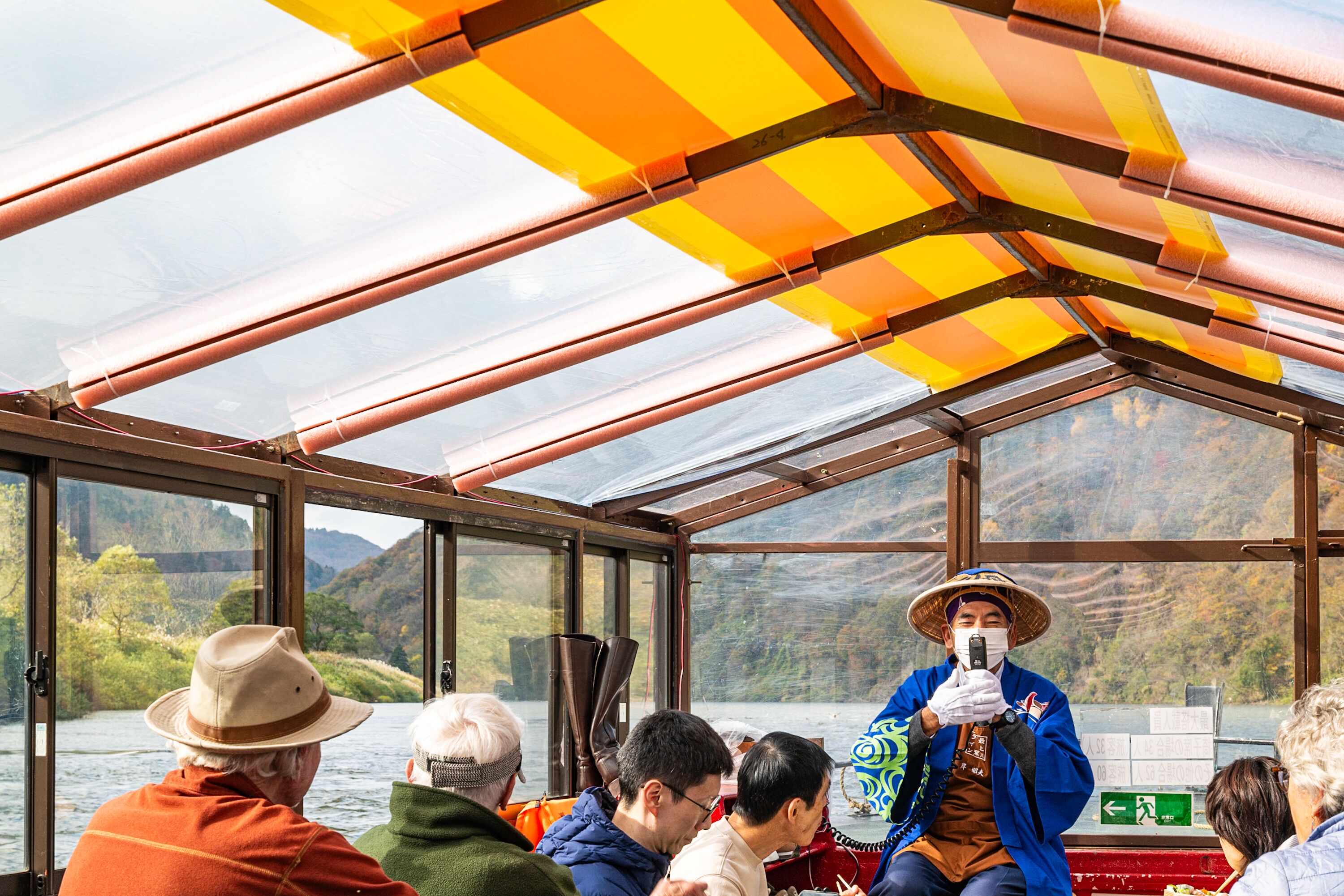 A boat with a glass roof and metal framework, passengers seated, and a guide in traditional attire.