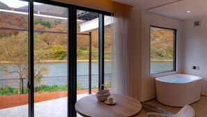 A modern bathroom with a large bathtub, a round table with a basket and a plate, and a view of a lake and autumn trees through large windows. - R MOGAMI (Tozawa)