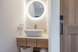 A modern bathroom with a round mirror, a white vessel sink, wooden cabinets, and a white basket.