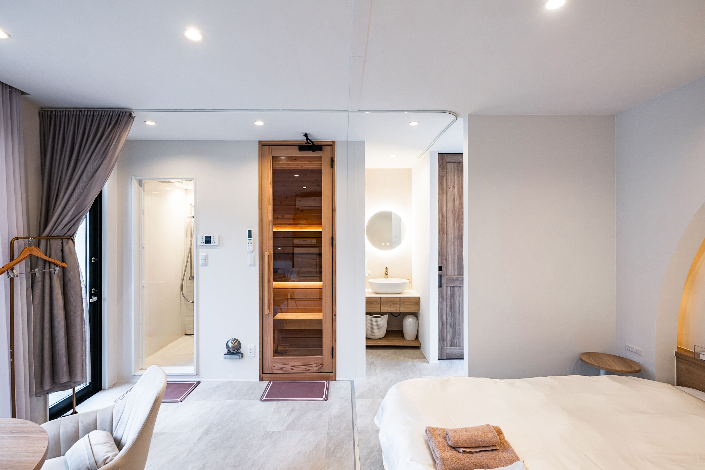 A modern hotel room with a bed, a seating area, a wooden wardrobe, and a bathroom visible through an open door.