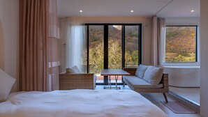 A modern hotel room with a large bed, a seating area with a table, and a view of autumn foliage through large windows. - R MOGAMI (Tozawa)