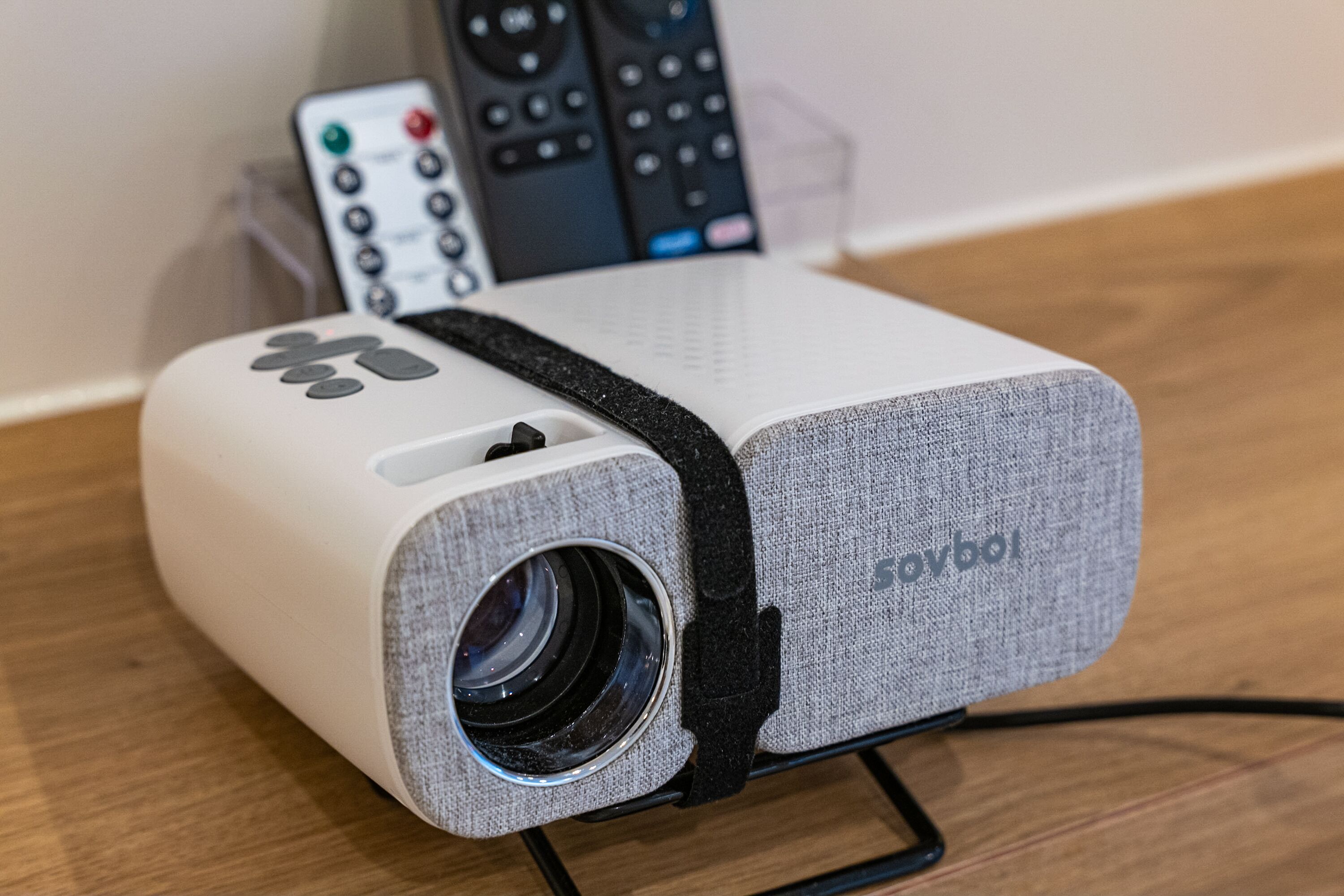 A portable projector with a lens, a remote control, and a power cord on a wooden surface.