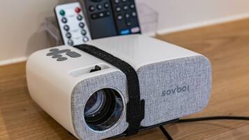 A portable projector with a lens, a remote control, and a power cord on a wooden surface.