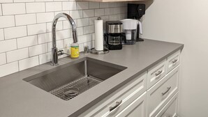 Une cuisine moderne dotée d’un évier à deux bacs, d’un robinet en acier inoxydable et d’une machine à café. Des étagères en bois, situées au-dessus de l’évier, servent à ranger des tasses et un moulin à café.
