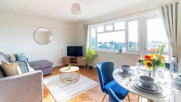 Una sala de estar moderna con un sofá, un televisor, una mesa de comedor para dos personas y un ventanal con vista a la ciudad.