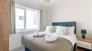 Una cama bien hecha con almohadas de diseño blanco y verde, una mesita de noche de madera con lámpara y una ventana con cortinas.