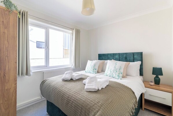 A neatly made bed with white and green patterned pillows, a wooden nightstand with a lamp, and a window with curtains. - Stunning Sea View 2 bedroom Sleeps 6- Free Parking (Bournemouth)