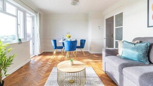 Una sala moderna con suelo de madera en patrón de espiga, un sofá gris con cojines azules, una mesa de centro redonda y una zona de comedor con sillas azules y una mesa blanca.